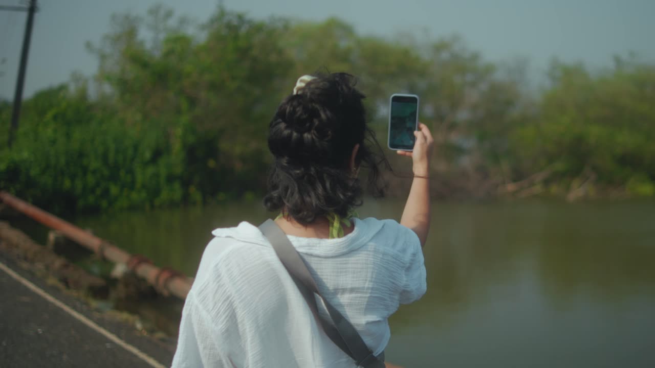 Uma jovem de camisa branca caminha por uma ponte panorâmica, usando o celular para registrar a exuberante paisagem do rio. Um momento de viagem autêntica e exploração moderna.