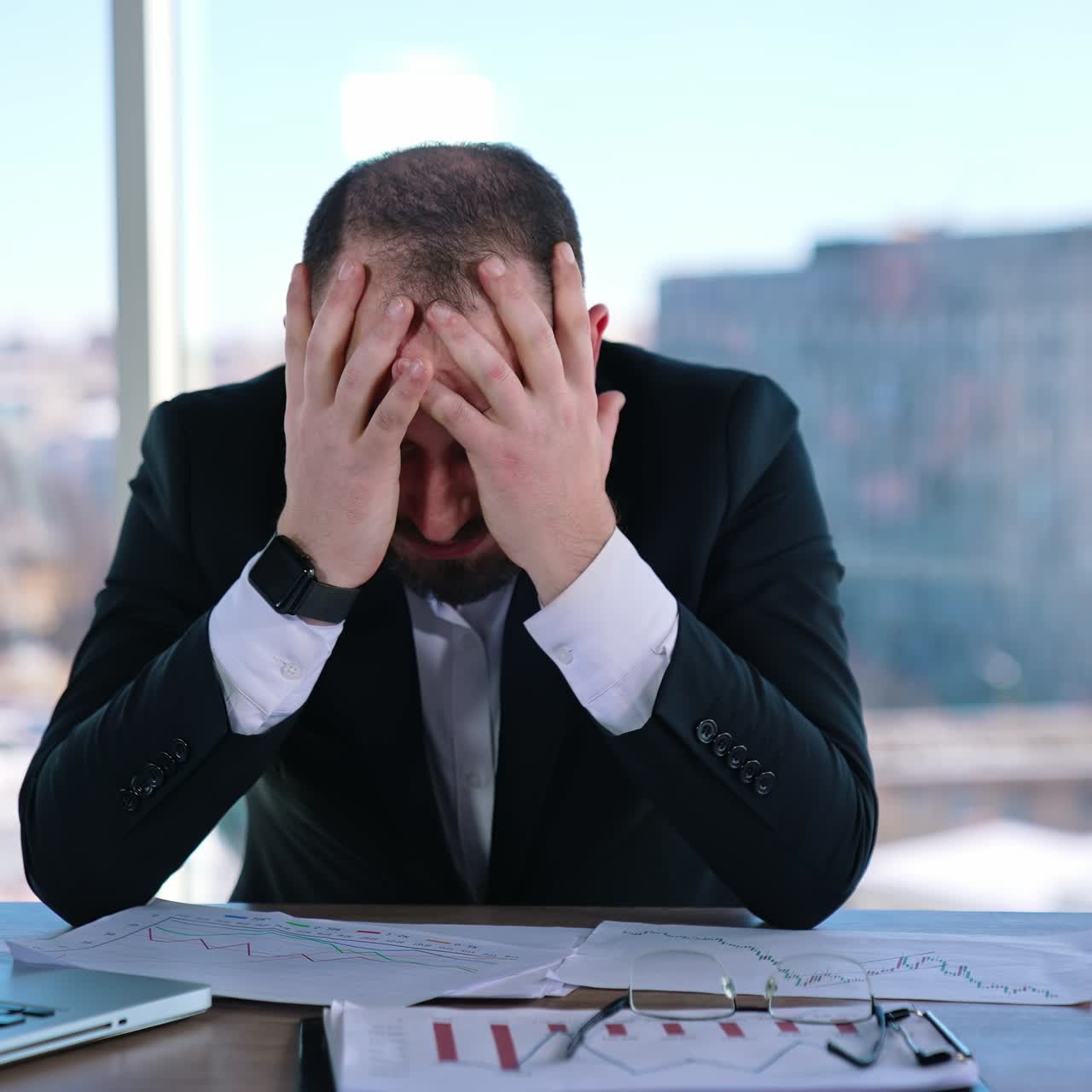 Portrait of a tired businessman at workplace. Bearded office worker in suit sitting at the table with a laptop on window background in the city