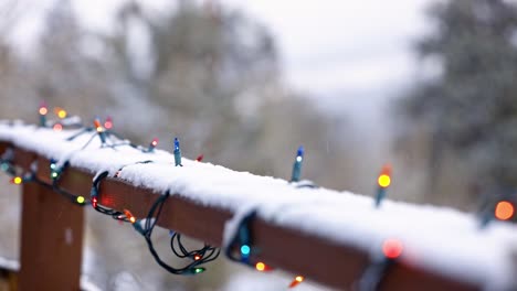 Close-shot-of-colored-Christmas-lights-on-a-wooden-railing-with-aspen-trees-in-the-background-and-snow-falling