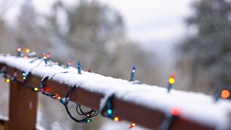 close shot of colored christmas lights on a wooden railing with aspen trees in the background and snow falling