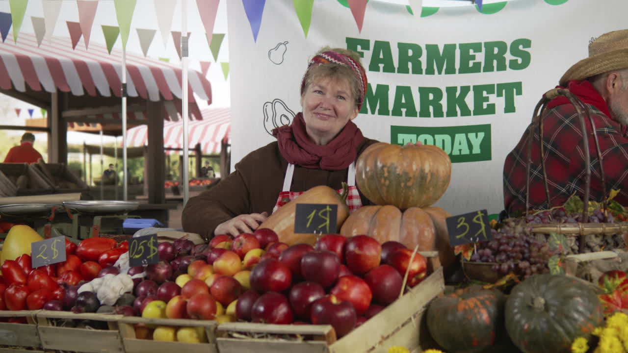 Woman Farmer Stands at the Stall and Smiles Woman Farmer Stands at the Stall with Fresh Colorful Fruits and Vegetables Looks at Camera Farmers Market Festival Vegetarian Organic and Healthy Food Agriculture