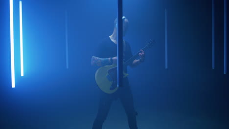 male guitarist playing song amidst blue lamps pan around view of bald man with headphones standing amidst bright blue lamps and playing electric guitar in dark room