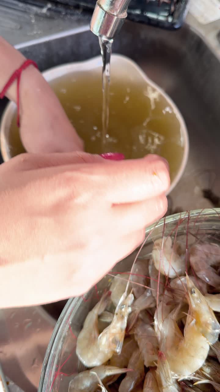 Manos de mujer limpiando camarones crudos bajo el grifo en una cocina doméstica, vista vertical para mariscos saludables y cocina.