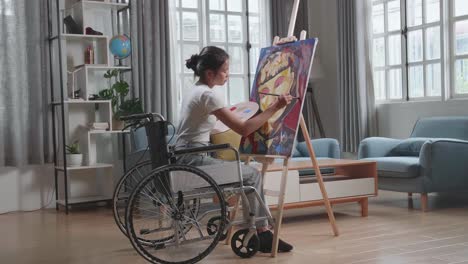 vista lateral de una artista asiática en silla de ruedas sosteniendo un pincel de colores mixtos pensando y pintando a una chica en el lienzo