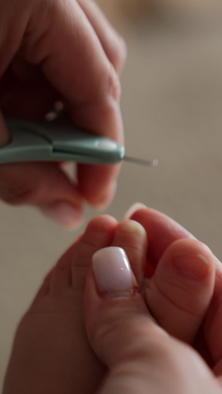 Mom's hands hold the tiny baby foot and using the little scissors to cut the toenails. Taking care of a child. Close up. Blurred backdrop. Vertical video