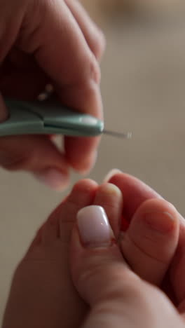 mom's hands hold the tiny baby foot and using the little scissors to cut the toenails. taking care of a child. close up. blurred backdrop. vertical video