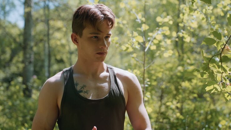 Front view of soldier looking forward while loosening headband and rolling fabric strap on hand, focusing ahead in sunlit forest clearing, sweat glistening on freckled skin under dappled light