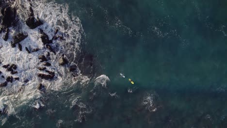 two surfers wating for waves near rocky coastline, top down zoom in drone view