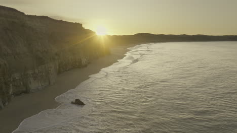 sunrise peaks behind fortrose cliffs with waves crashing on beach, aerial