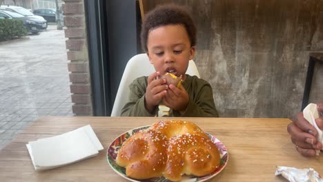 Adorable-and-exotic-three-year-old-black-child-enjoys-eating-a-Kinder-Surprise-seated-inside-a-cafeteria-next-to-his-mother