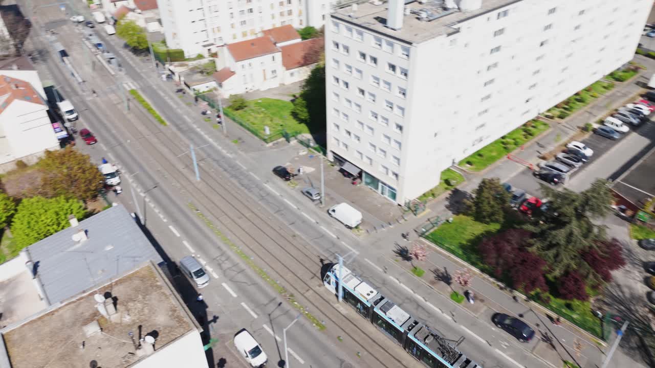 Drone top-down shot of a tram arriving at a station in suburban Paris, showing houses, buildings, and a sunny parking lot - France