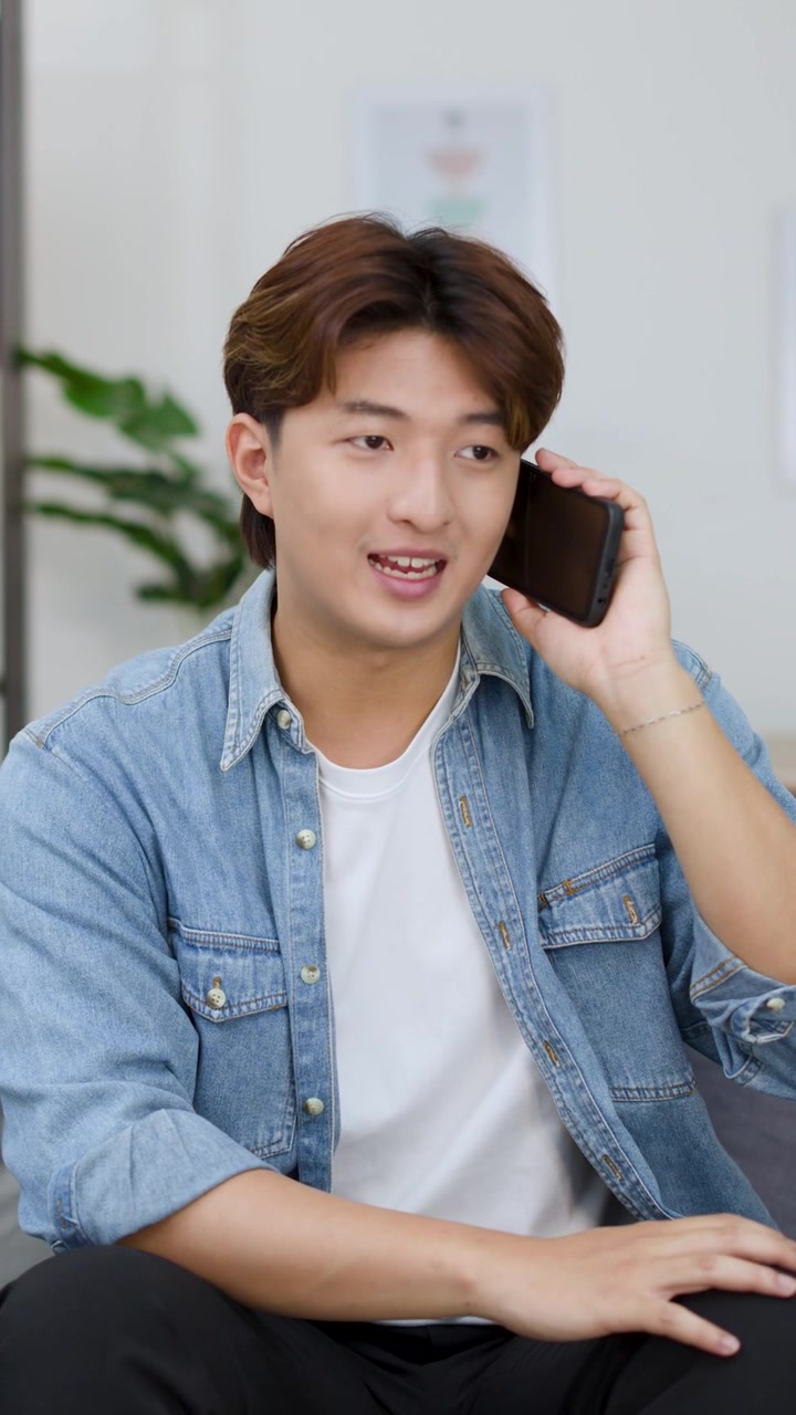 Un giovane uomo asiatico siede su un divano, compone un numero e parla al suo smartphone all'interno
