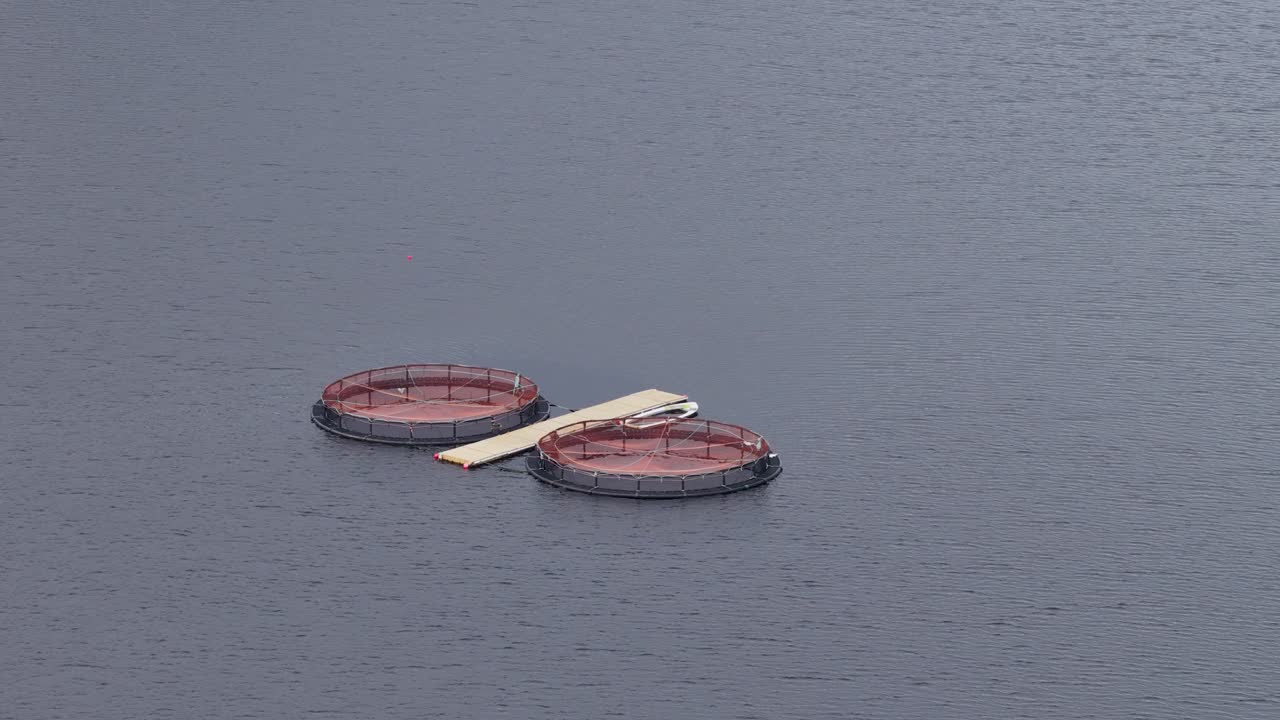 Two large circular fish farming tanks float on calm reservoir water, viewed from above in overcast daylight. Smooth aerial camera movement reveals isolated aquaculture structures