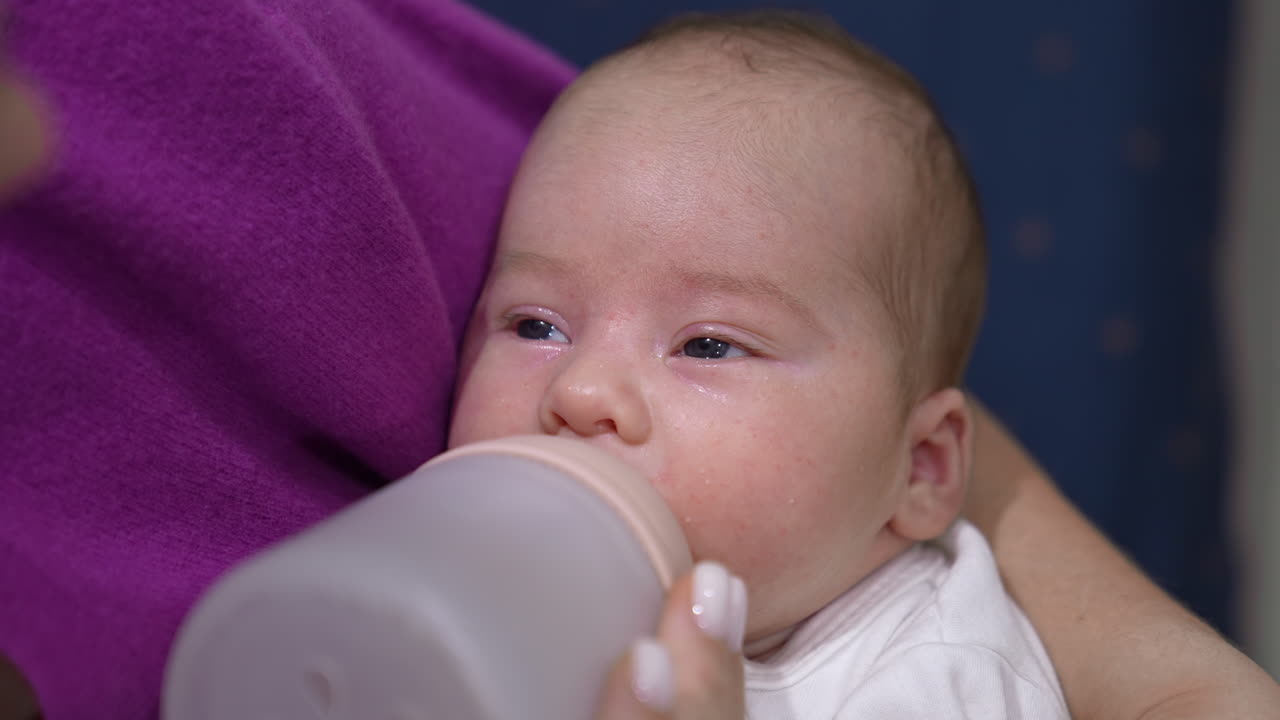 Cute infant suckling milk slowly from a bottle. Baby closing eyes and falling asleep while eating. Kid in mommy's hands close up.
