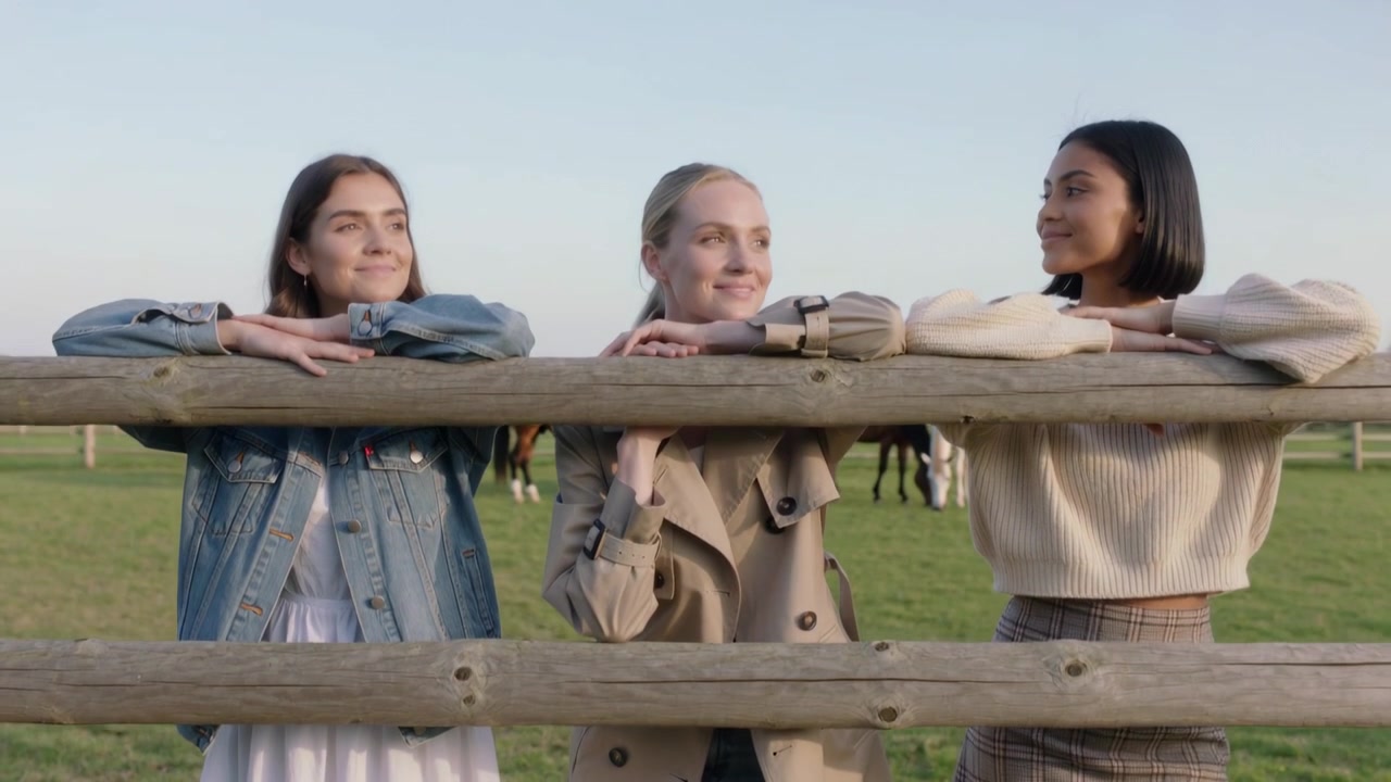 Drie vrouwen in spijkerbroek leunen tegen een weidehek, terwijl de camera terugtrekt en ze grazende paarden observeren.