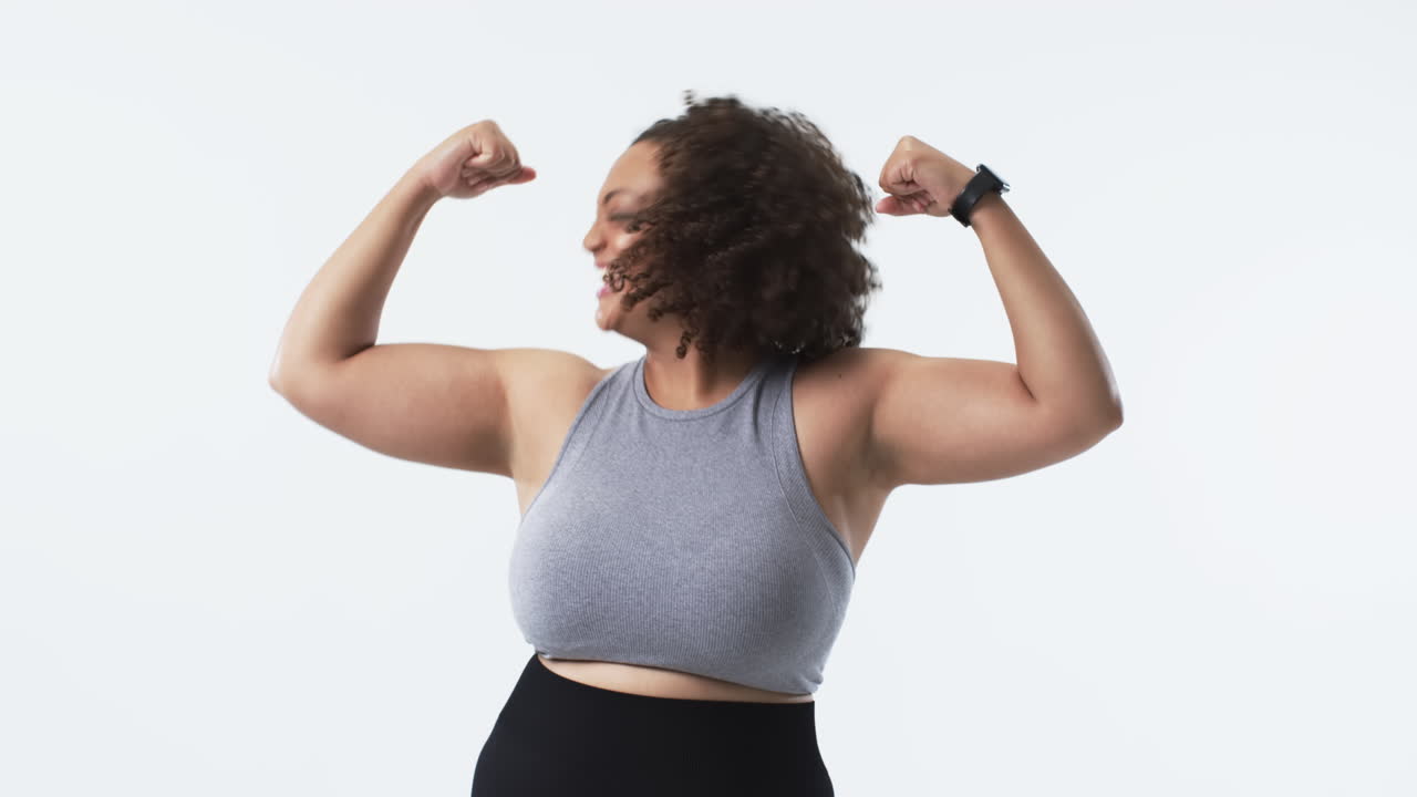 Young biracial plus size woman with curly hair smiling and flexing her arms in studio