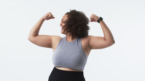 young biracial plus size woman with curly hair smiling and flexing her arms in studio