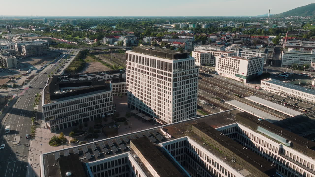 Drone moves toward the tall modern hotel, flying over the adjacent building and showing the railway tracks and a passing red train behind it