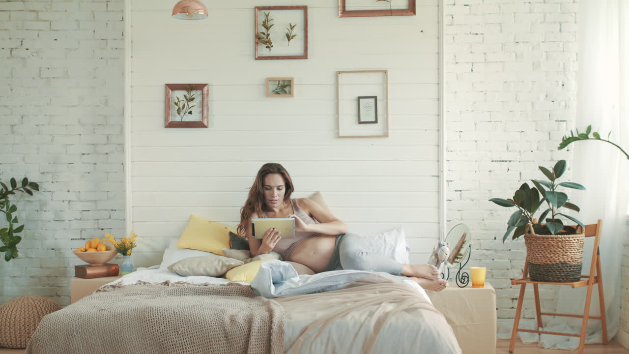 una embarazada feliz trabajando con una computadora portátil en la cama, una madre embarazada acariciando su vientre en casa.