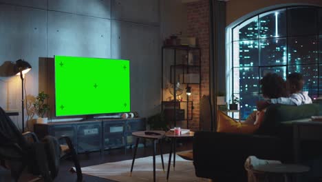 black couple in love sitting on a couch, watching green screen chroma key tv in modern apartment. boyfriend and girlfriend spend time together at home as a family . wide side back shot