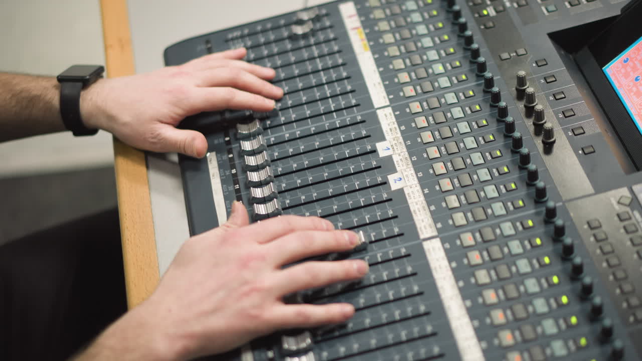 Close-up shot of hands adjusting audio faders on broadcast sound mixing desk. Professional audio mixing in control room with faders, dials, and sound equipment in use during media production