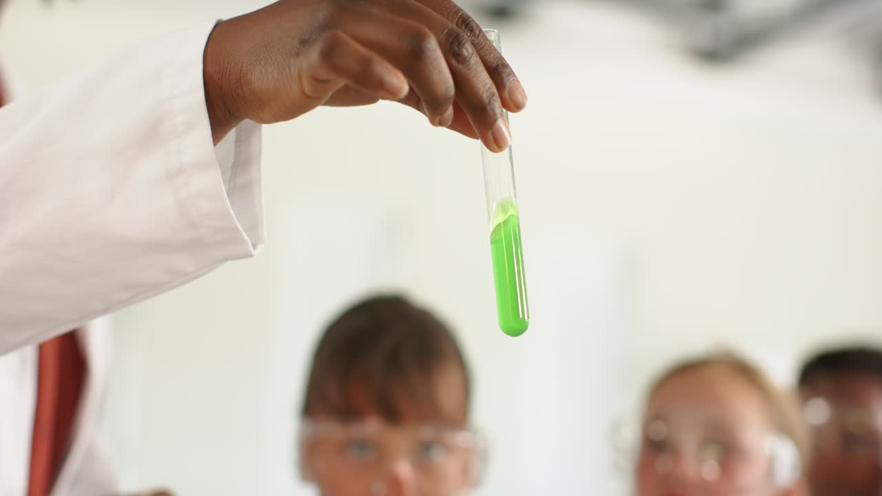 African American woman in lab coat leading demo, raising, tilting test tube green liquid for teens