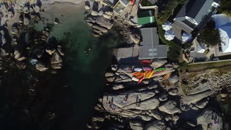 Birds-eye-drone-shot-of-Bakoven-beach-in-Camps-Bay,-Cape-Town---drone-is-ascending-over-some-stored-kayaks