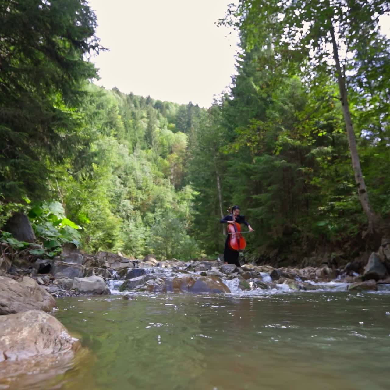 Cellist among mountain water. Professional female musician performing music with large musical instrument on green nature background