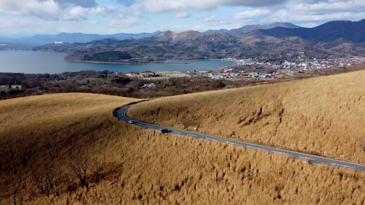 황금 잔디 언덕과 휘어지는 도로가 해안 마을로 이어집니다. 구름이 있는 파란 하늘 아래