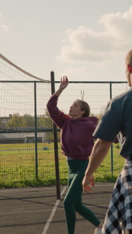 dama jugando al voleibol con el entrenador golpeando la pelota sobre la red mientras otros dos compañeros de equipo miran, con el campo de fútbol visible en el fondo y el cielo despejado sobre la cancha deportiva al aire libre