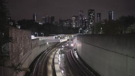 a dlr train station and train leaving in london during a clear night with buildings of canary wharf in the background