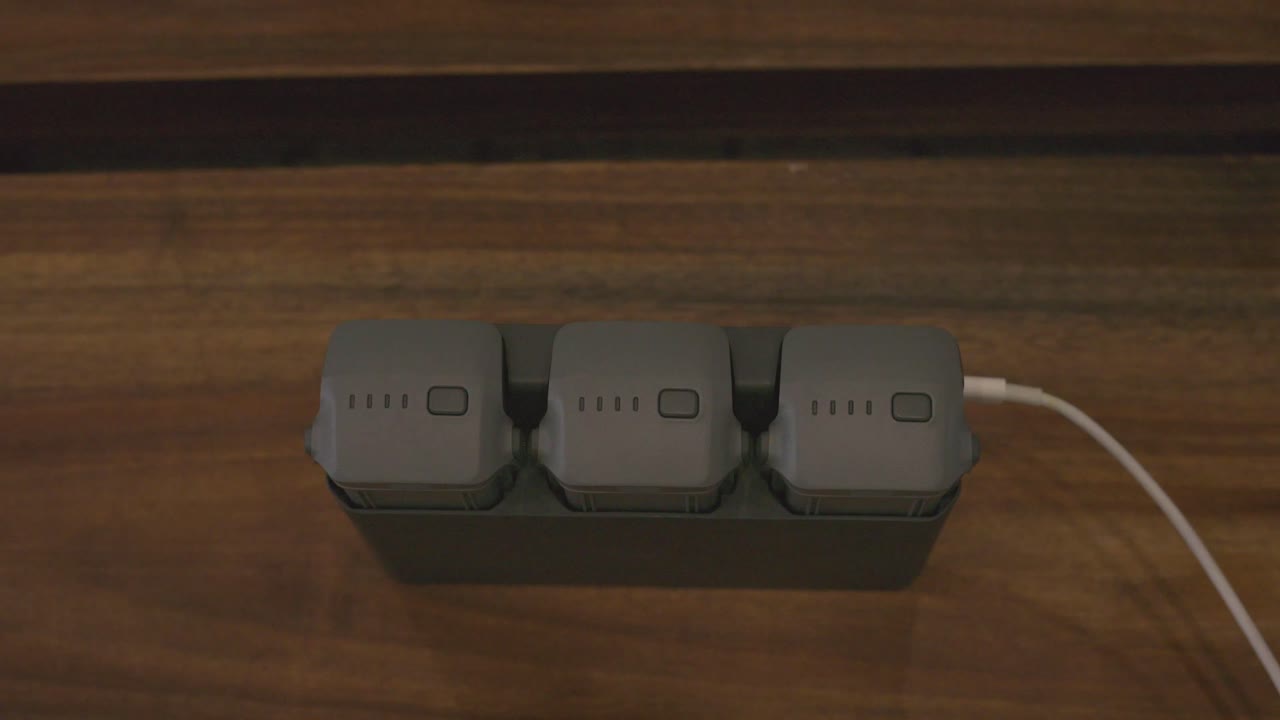 A set of three drone batteries placed in a charging dock on a wooden surface. One of the batteries shows illuminated green LED indicators, signaling an active charging process