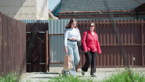three young ladies return from shopping, entering through open gate with happy expressions, one holding paper bag, casting shadows on paved path in sunny yard and brick building in background