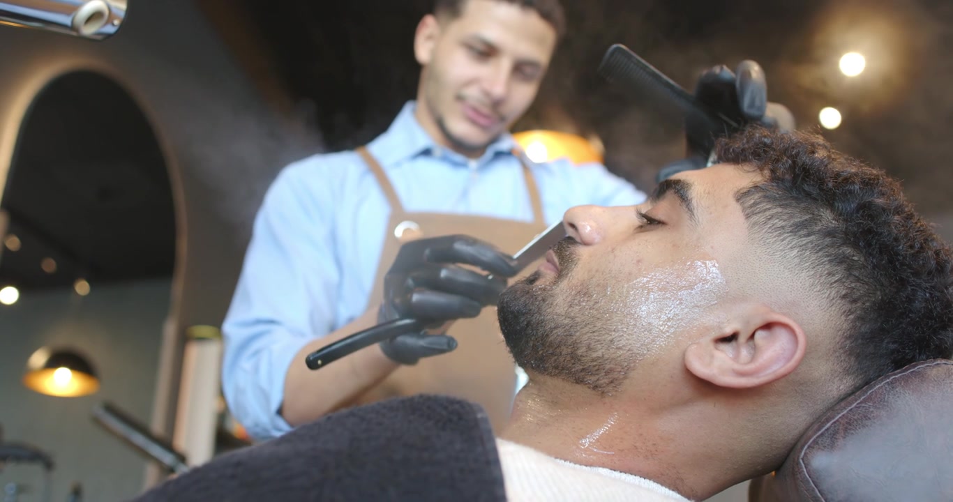 Barber i blå skjorte og sorte handsker damper og barberer en kunde i stolen, former skæg med en skraber, med plads til tekst.