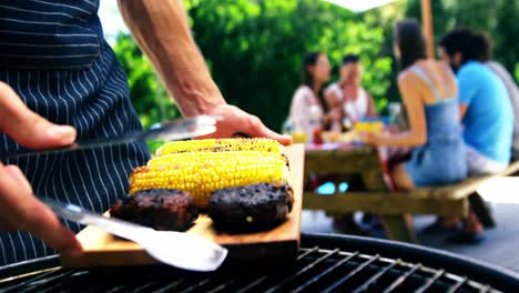 Man-putting-grilled-meat-and-corn-on-the-tray-for-serving-at-outdoors-barbecue-party