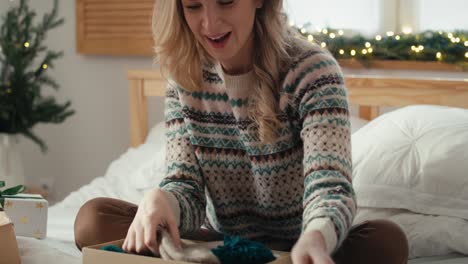 mujer caucásica sorprendida sentada en la cama y abriendo el regalo de navidad en el dormitorio