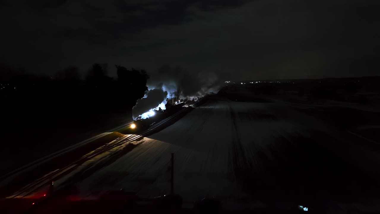 A steam locomotive moves steadily through a snow-covered area under the night sky, puffing clouds of steam. The soft glow of its headlights illuminates the surrounding landscape.