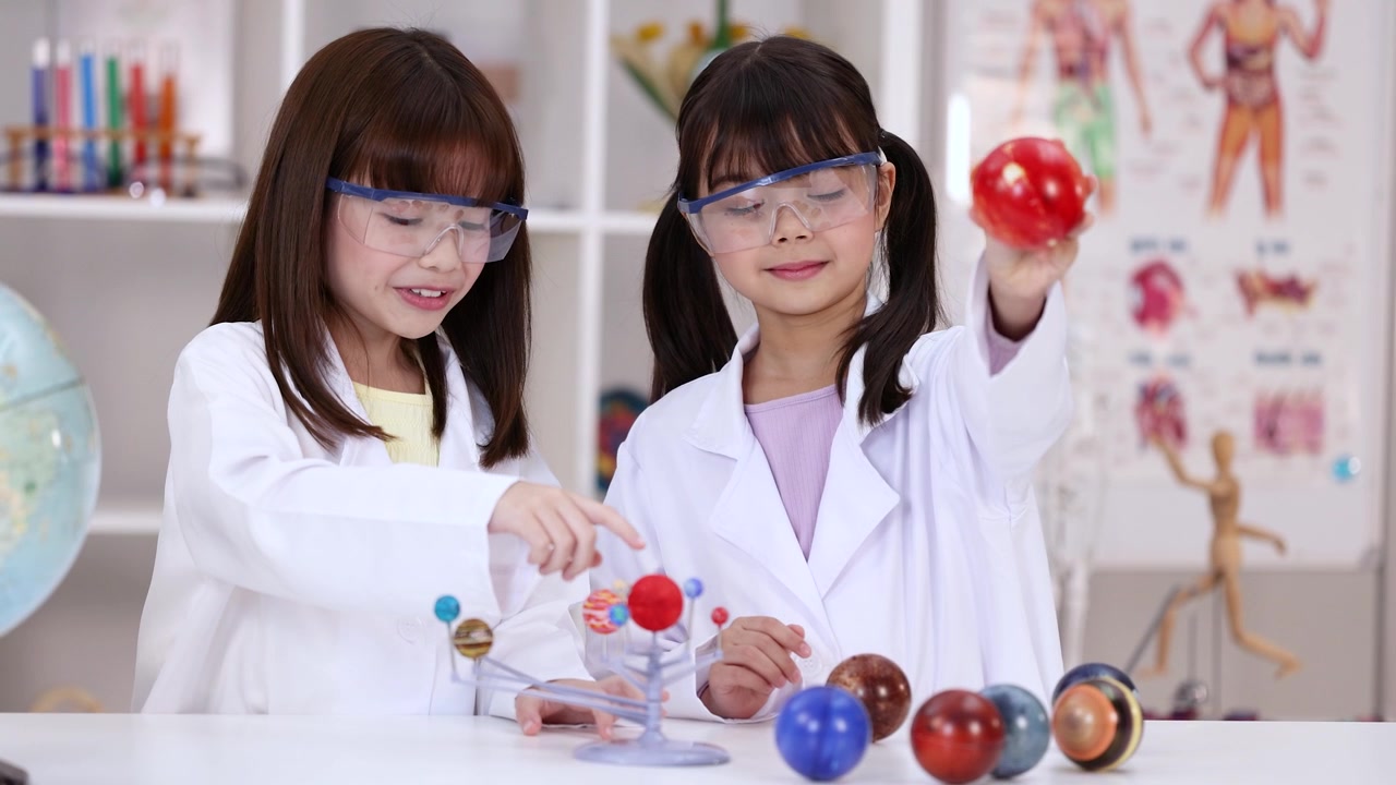 Two girls wearing safety goggles and lab coats examine a solar system model in classroom.