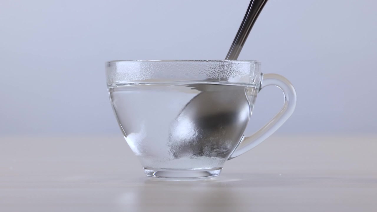 Una cuchara de metal revuelve polvo blanco en un vaso de agua sobre una mesa.