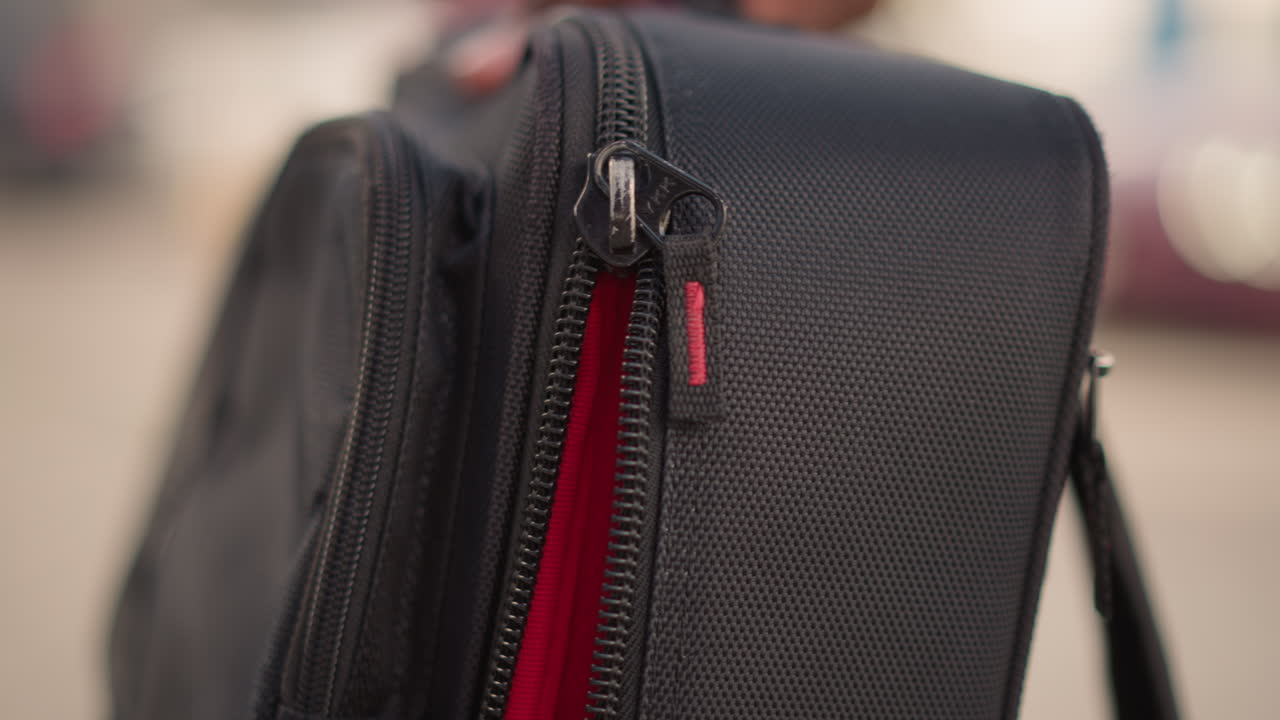 closeup black bag zipper red lining. tight shallow depth of field highlights textured synthetic fabric and metal slider as hand pulls zipper on urban sidewalk blurred cars and pedestrians suggest