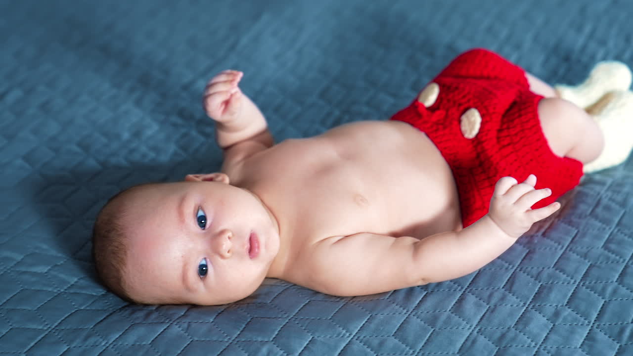 Blue-eyed Caucasian child lying on his back on the bed. Naked boy in red knitted shorts tossing little feet in white socks. Grey backdrop.