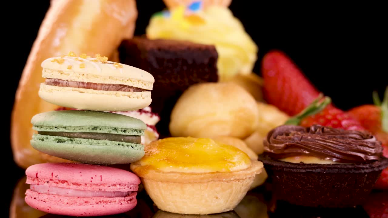 A rotating display of assorted bakery desserts including macarons, cupcakes, donuts, tarts, brownies, and strawberries, brightly lit against a black background with smooth camera movement