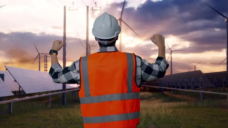 back view of a male engineer with safety helmet raising his hands celebrating while working with solar panel and wind turbines