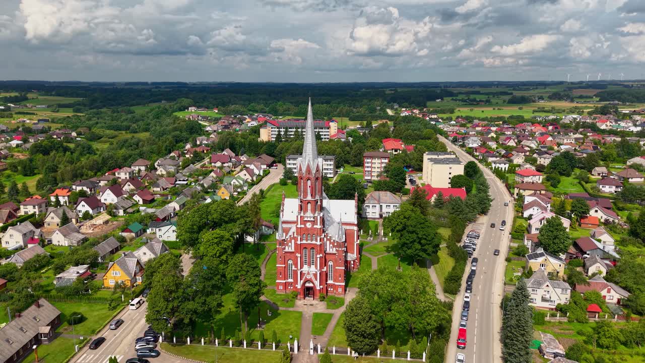 Aerial view of Šilalė St. Francis of Assisi Church surrounded by residential homes, green landscape, and scenic countryside in Lithuania under a dramatic cloudy sky