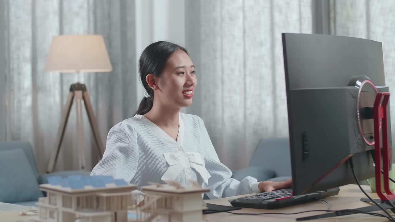 집 모델과 함께 미소 짓는 아시아 여성 엔지니어가 집에서 데스크에서 일하면서 머리를 긍정적으로 흔들고 있습니다.