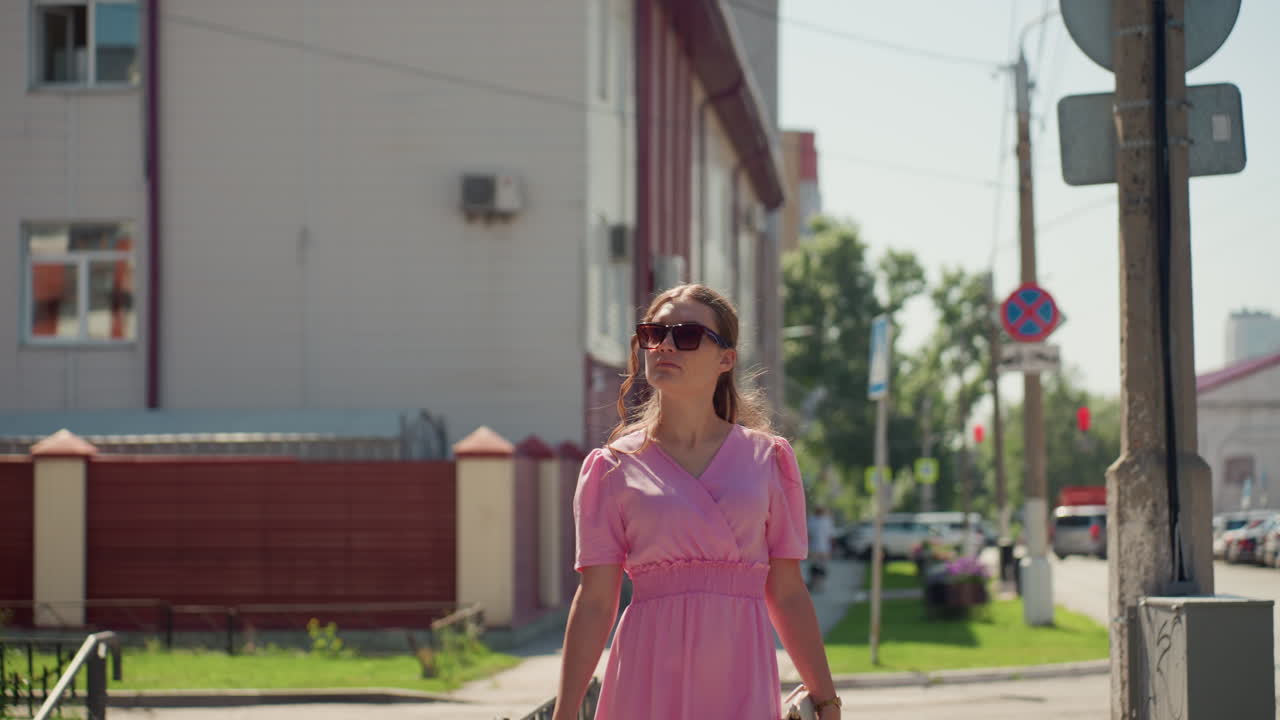 Kaukasische Frau mit rosa Kleid geht die Straße entlang, rückt ihre Sonnenbrille zurecht und schreitet selbstbewusst über den Fußgängerüberweg und an den Schaufenstern vorbei, Sommerliches Sonnenlicht, lässig-schickes Outfit, Lifestyle-Influencer-Stimmung, Nachbarschaft