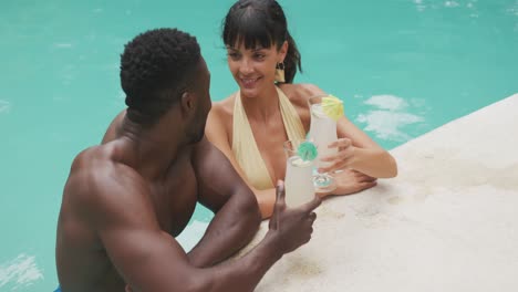 happy diverse couple wearing swimming suits and drinking drinks at swimming pool in garden