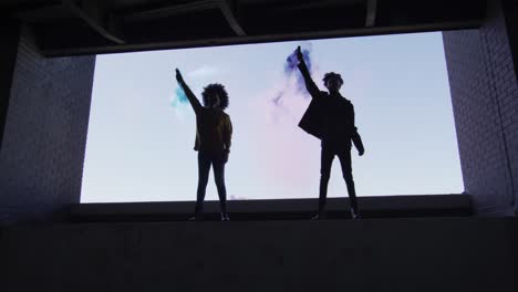 Mixed-race-man-and-woman-holding-blue-and-purple-flares-standing-in-an-empty-building