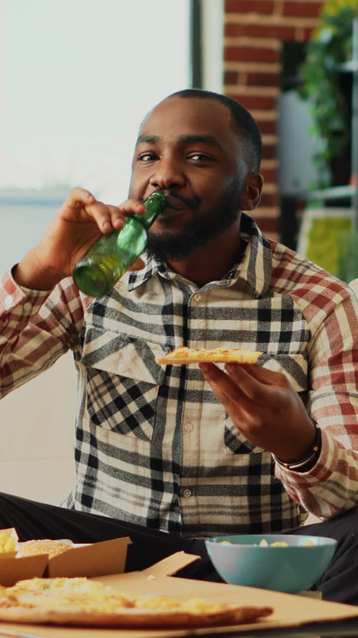 Vertical video: African american man eating slice of pizza at home
