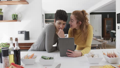 Feliz-Pareja-De-Lesbianas-Caucásicas-Paradas-En-El-Mostrador,-Hablando-Y-Usando-Una-Tableta-En-Una-Cocina-Soleada